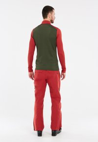Rood shirt met lange mouwen met een groene achterkant, gecombineerd met rode waterdichte broek met ritszakken en flared manchetten. Zwarte laarzen zichtbaar.