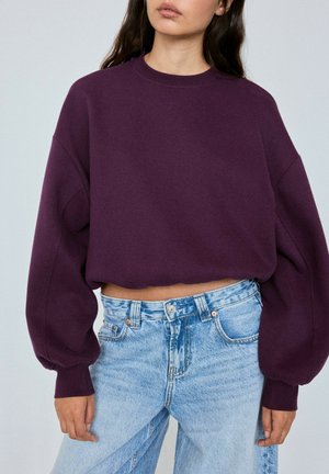 Femme portant un sweat violet ample et court et un jean bleu clair taille haute sur un fond uni.
