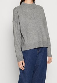 Pull en maille gris avec un col roulé, des épaules tombantes, des poignets et un ourlet côtes. Porté sur un pantalon en denim bleu ample.