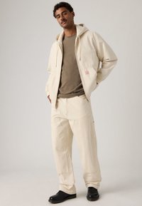 Μπεζ φούτερ με ρίγες και αντίστοιχο παντελόνι φτιαγμένα από υφασμάτινη υφή. Το φούτερ έχει κορδόνι και μια μικρή λεπτομέρεια με λογότυπο στην τσέπη.