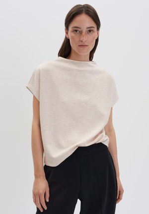 Femme aux longs cheveux bruns portant un haut beige à manches courtes et un pantalon noir, se tenant devant un fond clair uniforme.