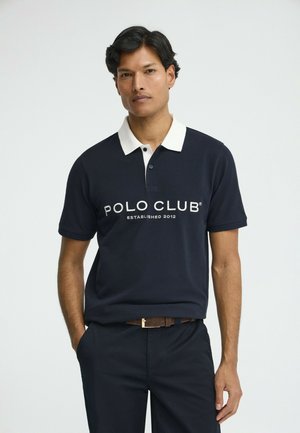 Uomo che indossa una polo blu navy con colletto bianco e la scritta "POLO CLUB Established 2012", abbinata a pantaloni scuri e cintura marrone.