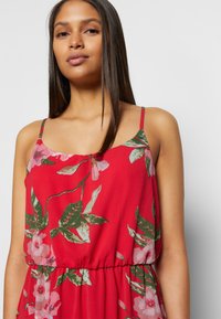Vestido floral vermelho com alças finas, apresentando padrões de flores em rosa e verde, tecido leve e uma cintura elástica para um ajuste confortável.