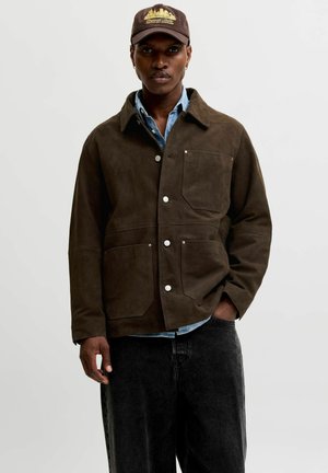 Homme portant une veste en daim marron à boutons, une chemise en denim bleu clair, un jean noir et une casquette marron foncé avec une broderie de la silhouette de la ville.