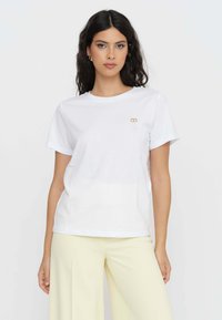 T-shirt bianco in cotone con maniche corte, scollatura rotonda e un piccolo emblema dorato sul petto. Tessuto liscio con una vestibilità comoda.