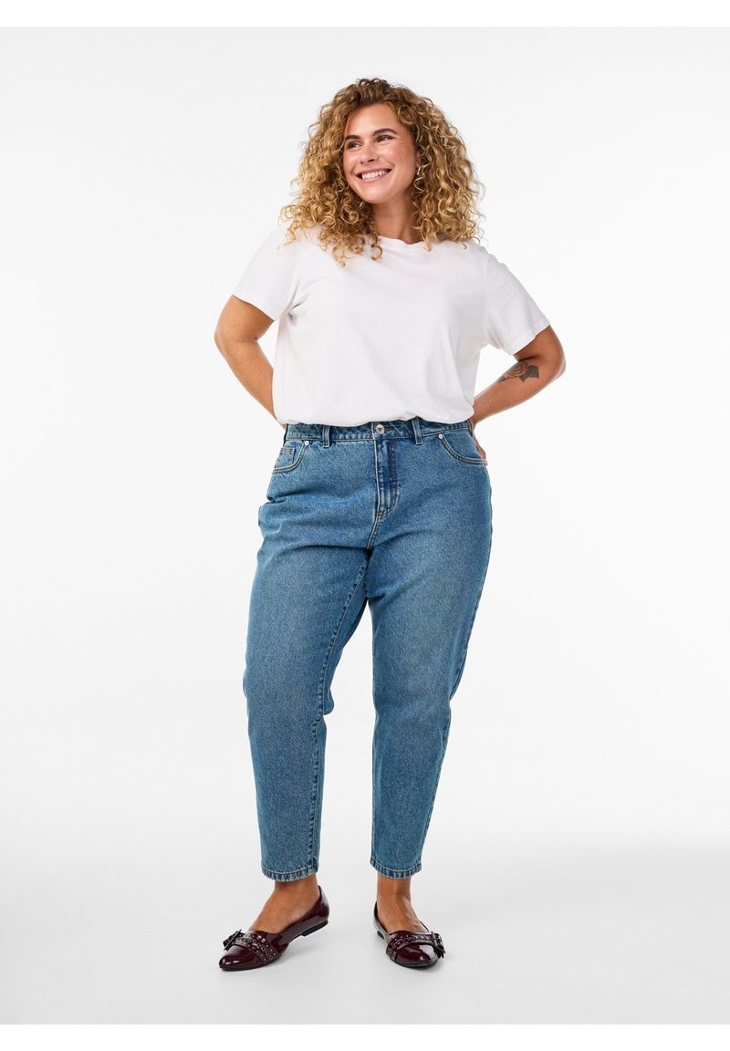 Mujer sonriente con el pelo rizado, de pie con las manos detrás de la espalda, lleva una camiseta blanca, vaqueros azules y zapatos planos rojo oscuro sobre un fondo blanco liso.