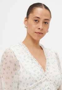 Vit blus med blommönster och ljusblå och gröna accenter. Har volangkanter och genomskinliga puffärmar. Slät textur och en delikat tryck.
