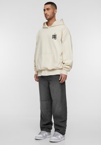 Sweat à capuche crème avec un logo noir, poche kangourou et coupe décontractée, associé à un jean gris foncé ample et des baskets blanches.