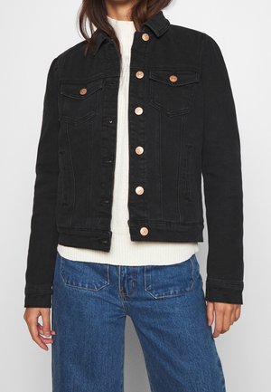 Kvinde iført en sort knappet denimjakke over en hvid strikket sweater og blå jeans, stående mod en ensfarvet baggrund.