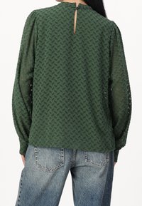 Blouse verte à motifs avec des manches longues, dotée d'une texture en chevrons et d'une petite fermeture à bouton à l'arrière, portée sur un jean bleu clair.