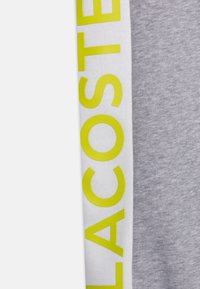 Šedá tkanina se hladkým povrchem, na které je bílý pruh s velkým, jasně žlutým nápisem "LACOSTE".