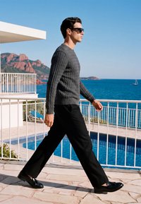 Homme portant des lunettes de soleil, un pull gris et un pantalon noir marchant sur la terrasse près de la piscine avec l'océan et des collines rocheuses en arrière-plan par une journée ensoleillée.