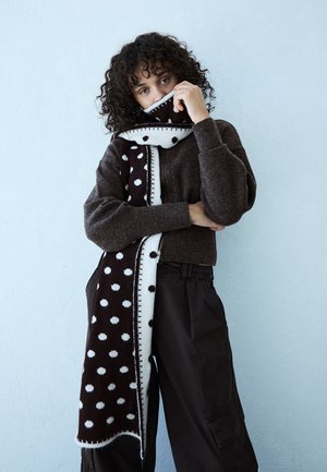 Personne aux cheveux bouclés portant un pull marron foncé et un pantalon large, couvrant partiellement son visage avec une longue écharpe noire et blanche à pois.