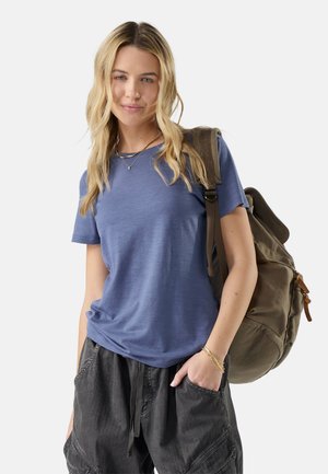 Young woman with long blonde hair wearing a blue t-shirt and black pants, carrying a large brown backpack over one shoulder.
