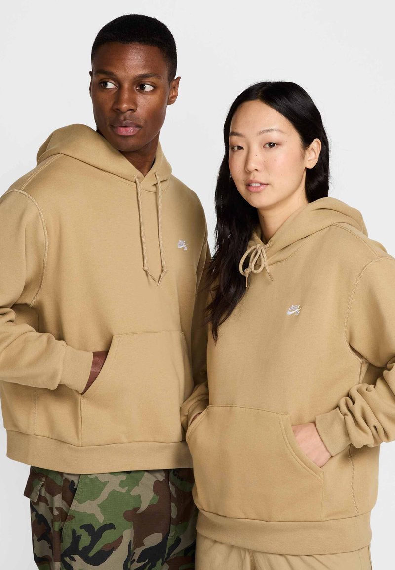 To personer iført store, beige hettegensere med frontlomme og snor på hetten. Hettegenserne har en liten hvit logo på brystet.