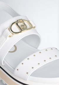 Sandal bianca con finitura lucida, dotata di una fibbia in metallo dorato, cinturino testurizzato e decorazioni di piccoli borchiette metallici sulla parte superiore.