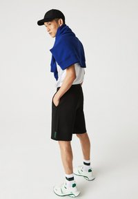 Shorts noirs avec poches latérales, accompagnés d'un pull bleu drapé sur les épaules, et d'un t-shirt blanc à motifs. Baskets blanches avec accents verts.