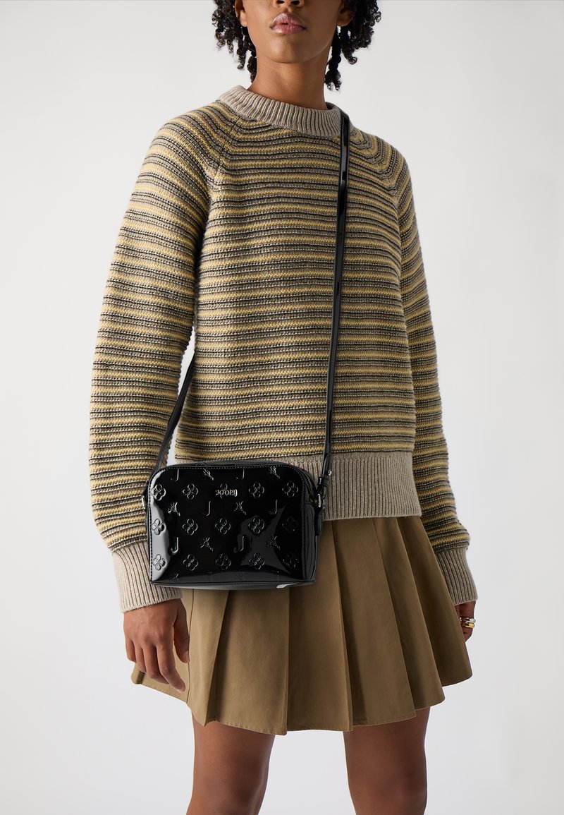 Borsa a tracolla in pelle verniciata nera con loghi embossati; forma rettangolare, cinturino regolabile e accessori lucidi; indossata con un maglione a righe.