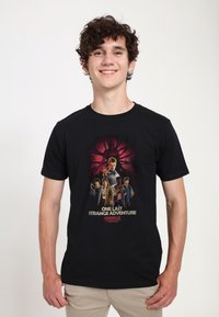 Schwarzes Baumwoll-T-Shirt mit einem Grafikdruck von Figuren aus einer TV-Show, unten mit dem Text "ONE LAST STRANGE ADVENTURE".