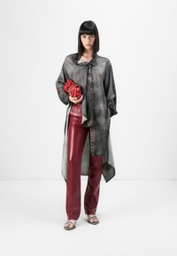 Pilka, per didelį dydį shirt su kaklaraiščio uždarymu, derinama su blizgiomis, burgundiško raudonumo dirbtinės odos kelnėmis. Modelis laiko raudoną rankinę, stovėdama ant paprasto fono.