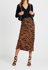 Schwarzes V-Ausschnitt-Oberteil kombiniert mit einem langen, braun-orange gestreiften Rock im Tigerdesign. Das Model trägt schwarze, Riemchen-Absätze und präsentiert eine glatte Stofftextur.