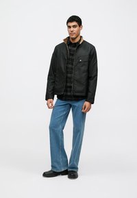 Black bomber jacket with a brown collar, chest pocket, worn over a black plaid shirt, paired with blue jeans and black footwear.