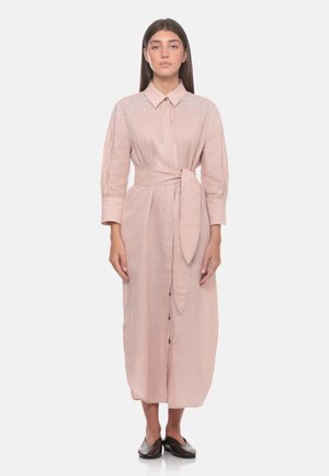 Donna che indossa un lungo vestito rosa chiaro con bottoni, cintura annodata in vita e spalle decorate, in piedi rivolta verso il davanti con espressione neutra.