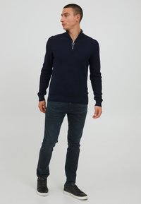 Vīrietis valkā tumši zilu zip-up džemperi, tumšas džinsus un melnas kedas, stāv un skatās pa kreisi uz pelēka fona.