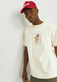 Cremefarvet T-shirt med korte ærmer, der har et trykt design af hænder i jordnære toner, samt en rød kasket med hvid tekst.