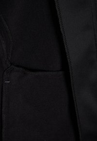 Nahaufnahme von schwarzem Fleece-Stoff mit detaillierter Naht und einer kleinen rechteckigen Tasche auf der linken Seite.