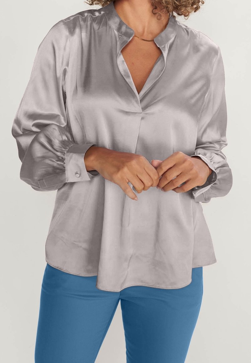 Femme portant un chemisier en satin argenté à manches longues avec un col fendu et un pantalon bleu, mains lâchement jointes à la taille.