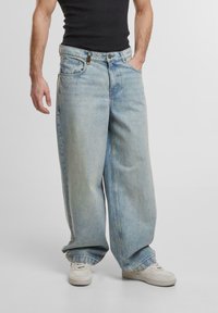 Pantaloni in denim oversize blu chiaro con design a gamba dritta, dotati di cinque tasche e dettagli di cucitura visibili. Indossati con una canotta nera.