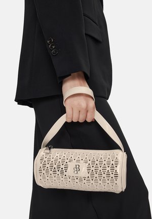Persona vestida con ropa negra sosteniendo un bolso cilíndrico beige con un patrón de recortes geométricos y una correa para la muñeca.