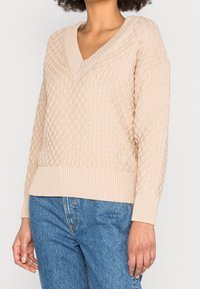 Una donna che indossa un maglione a V in maglia testurizzata beige abbinato a jeans in denim blu, in piedi con una mano rilassata al suo fianco.