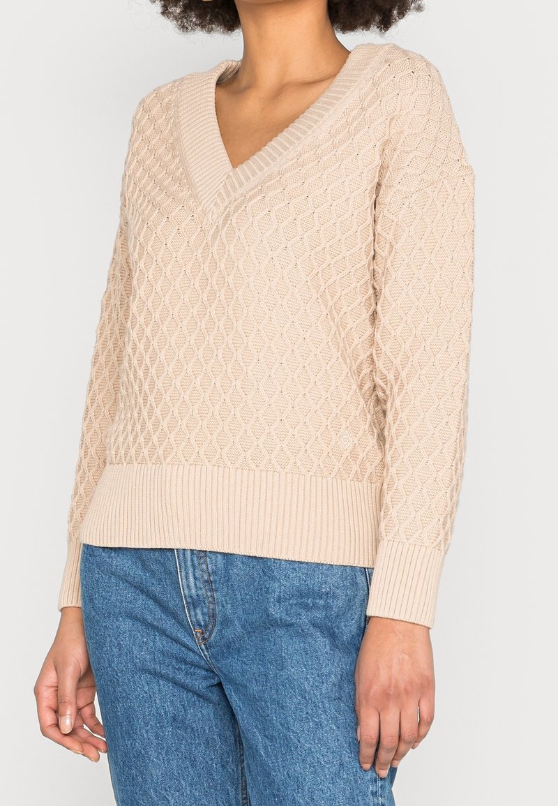 Una donna che indossa un maglione a V in maglia testurizzata beige abbinato a jeans in denim blu, in piedi con una mano rilassata al suo fianco.