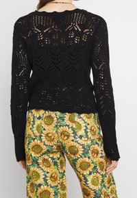 Suéter negro de punto con patrón de calado, mangas largas y dobladillo acanalado, combinado con pantalones florales con estampado de girasoles y detalles en verde.