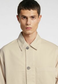Beżowa bawełniana koszula-kurtka z ostrym kołnierzem, dwoma guzikami, kieszenią na piersi i gładką fakturą.