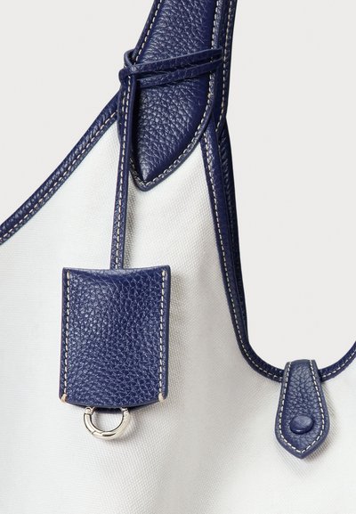 Λευκή τσάντα από καμβά με επένδυση από δέρμα σε σκούρο μπλε χρώμα, ραμμένα άκρα και μικρή δερμάτινη ετικέτα με ασημένιο μεταλλικό κρίκο προσαρτημένο στο λουρί.