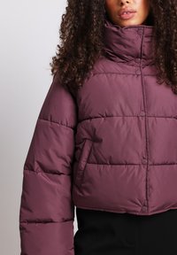 Plumón en color malva oscuro con cuello alto, diseño acolchado y botones a presión. Presenta bolsillos laterales y una silueta corta.