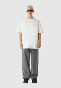 Bela kratka majica s kratkimi rokavi z vijoličnim logotipom, sivi ohlapni jeans in črne superge. Model nosi črne sončna očala. Enostavna ozadja.