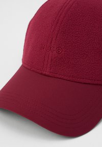 Rode pet van zacht fleece met een gebogen rand, voorzien van het geborduurde "HUGO" logo aan de voorkant en gestikte details langs de rand.