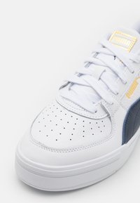 Baskets blanches Puma avec lacets blancs, bout perforé, bande latérale marine et logo Puma doré sur la languette et le côté.