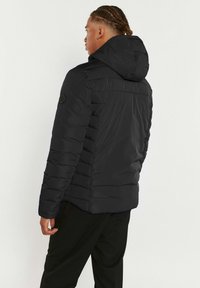 Doudoune noire avec capuche, présentant une texture matelassée, un design ton sur ton et un badge sur l’épaule gauche. Coupe ajustée, coutures visibles.