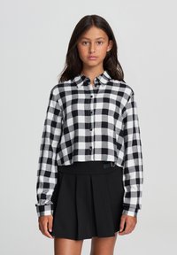 Camicia a maniche lunghe cropped in un motivo a scacchi bianco e nero, con bottoni, colletto e un tessuto morbido. Abbinata a una gonna plissettata nera.