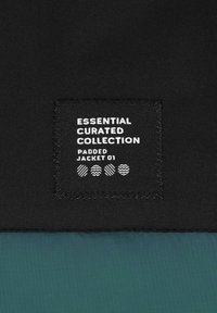 Zwarte stof met een rechthoekig label waarop staat "Essential Curated Collection, Padded Jacket 01." De tekst heeft witte en gemusterde accenten.