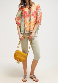 Helle, monivärinen kukkabloose leveillä hihoilla, sidottu kaula-aukko; vaaleanvihreät vajaamittaiset housut; keltainen olkakassi, jossa on tupsu; helmillä varustetut sandaalit.