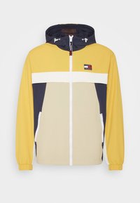 La giacca a vento presenta blocchi di colore giallo, beige e blu navy, con una striscia bianca, una chiusura con zip e tasche laterali. Cappuccio e polsini regolabili.
