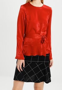 Top lungo a maniche con una cintura in vita di raso rosso, abbinato a una gonna nera con un motivo a griglia bianca. Tessuto liscio e design aderente.