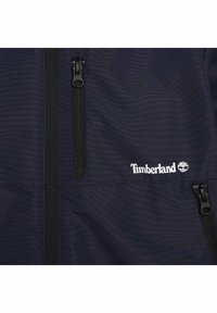 Námořnická bunda se dvěma černými zipy, texturovaná látka a bílé logo Timberland. Má strukturovaný design a praktické kapsy na zip.