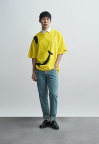 T-shirt oversize jaune avec un motif de smiley noir, associée à un jean bleu clair et des chaussures noires. Détail de col blanc visible.
